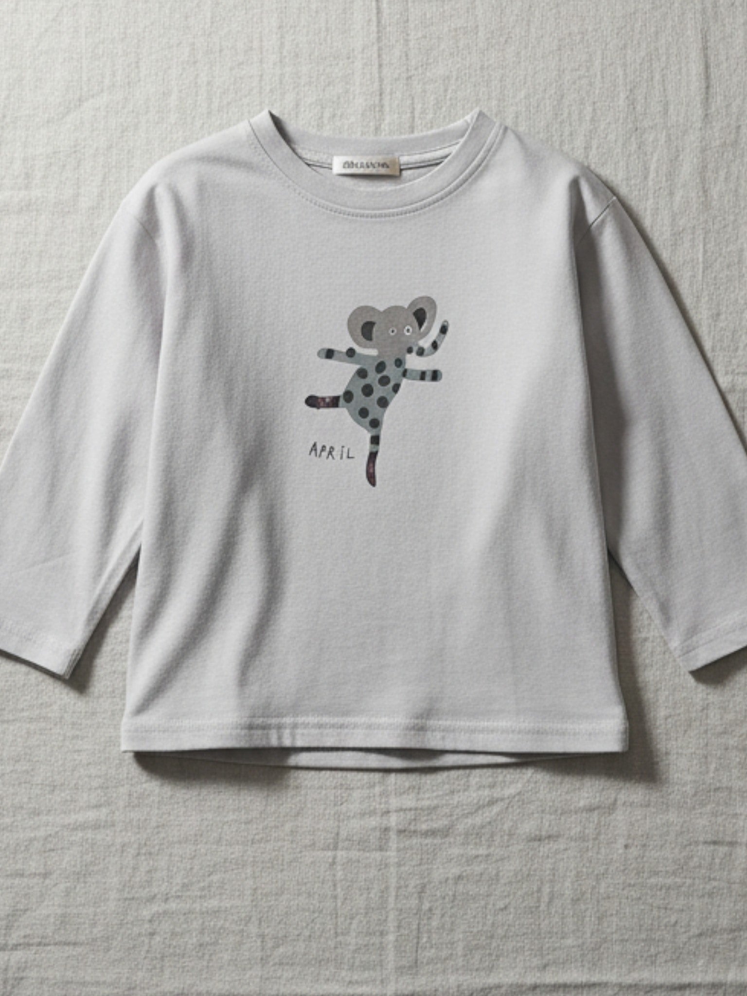 Children's light gray long-sleeve t-shirt featuring a playful spotted elephant illustration with the text "APRIL" printed below.