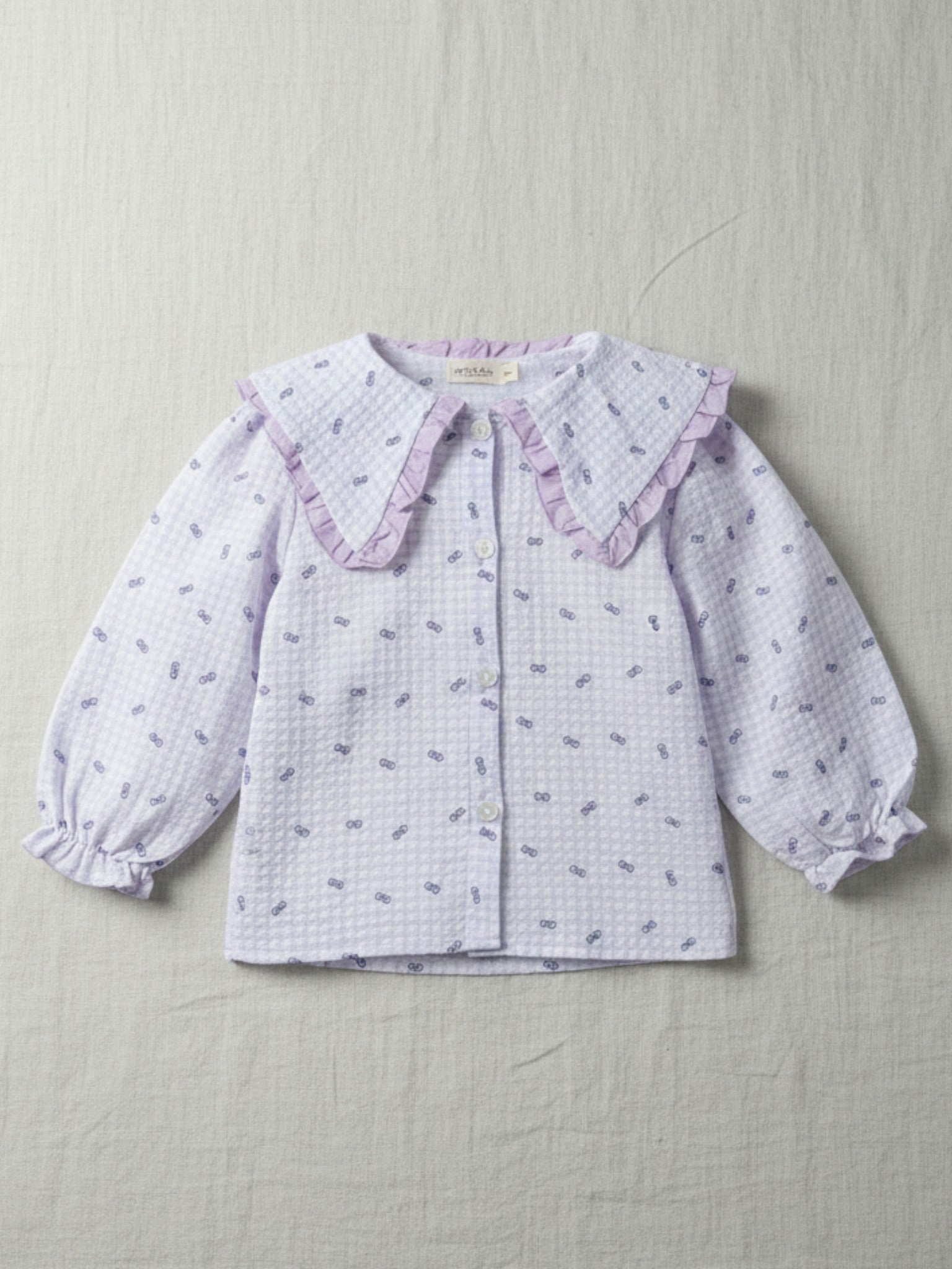 Girls' lilac and white seersucker blouse featuring a small bow print, a large purple-frilled Peter Pan collar, and elasticated ruffle cuffs