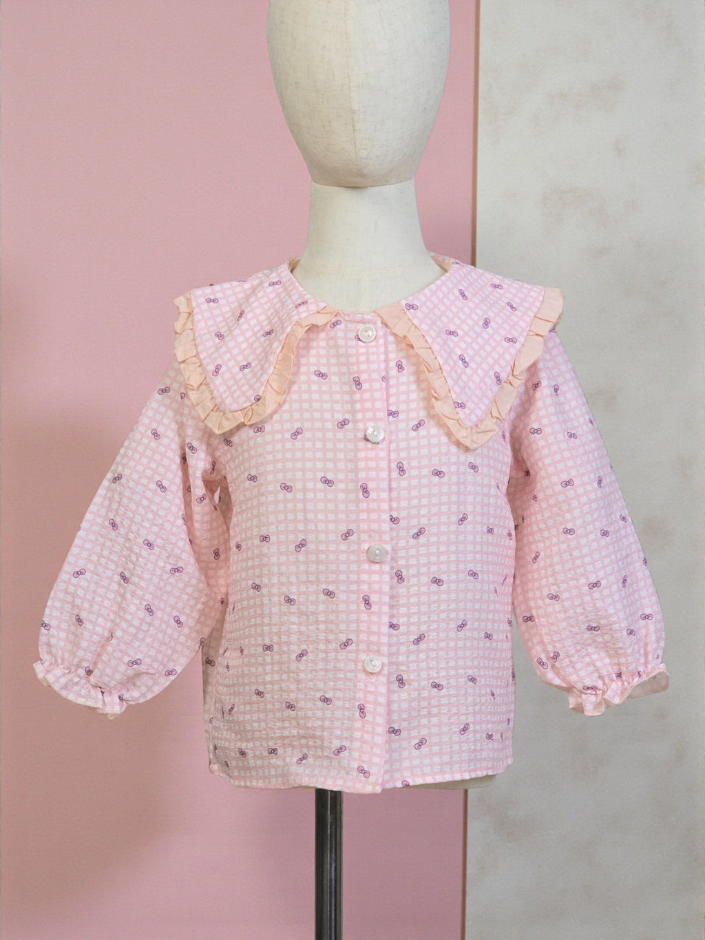 Pink checkered blouse with ruffled collar on a mannequin against a pink and gray background. Pink