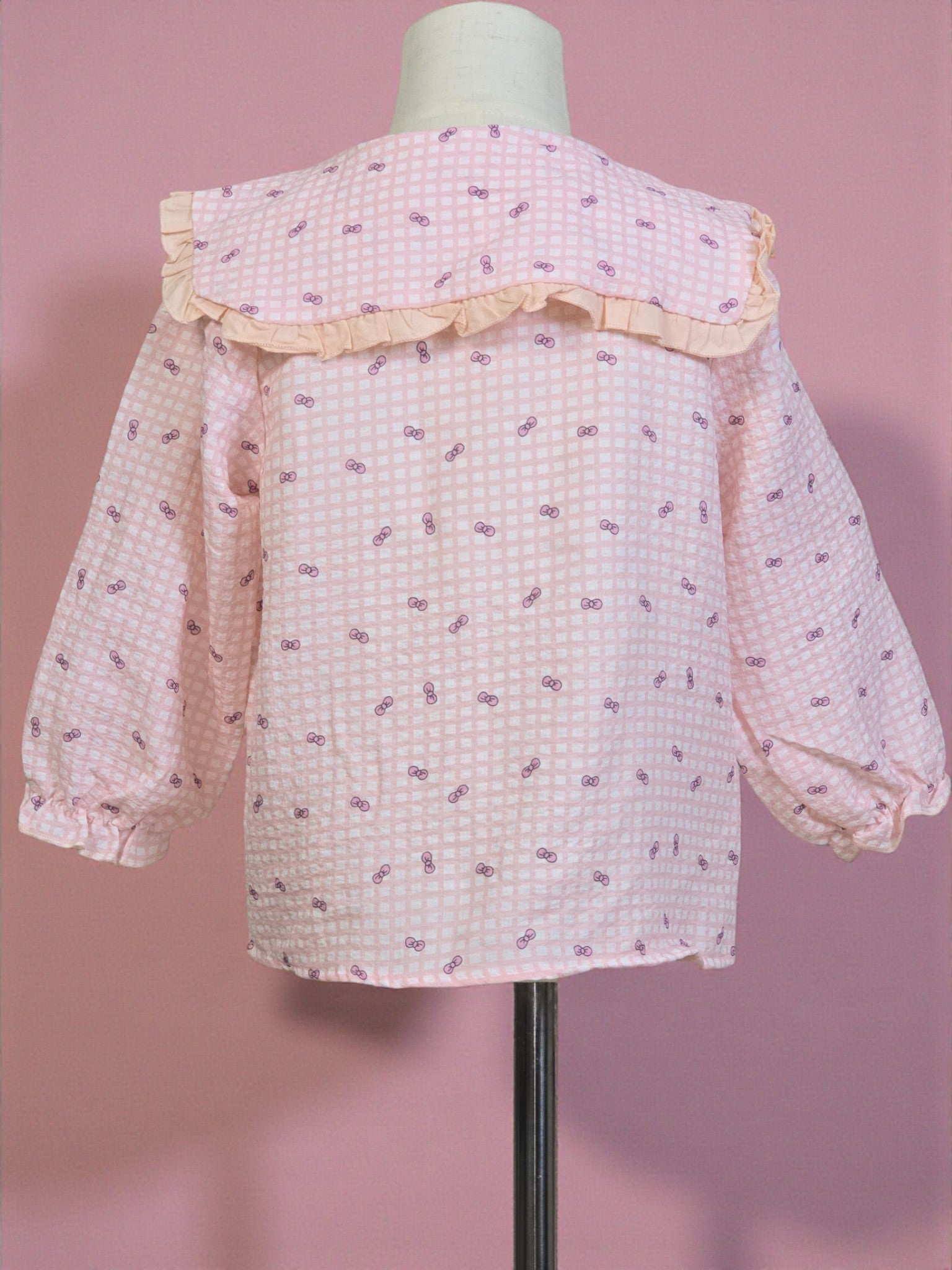Pink checkered blouse with ruffled collar on a mannequin against a pink background. Pink