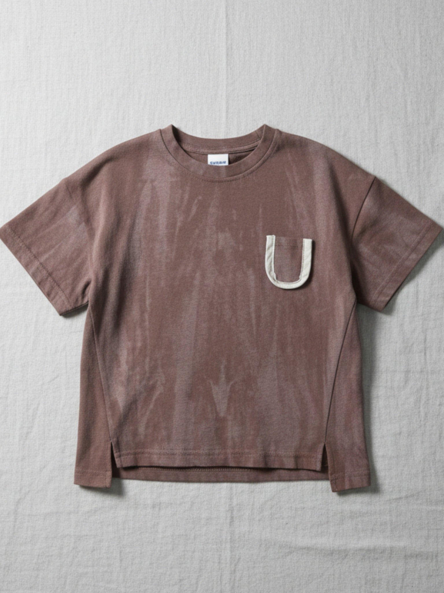 Children's short-sleeve t-shirt in a faded, washed brown finish featuring a white contrast pocket with a brown letter "U" and side slits at the hem.