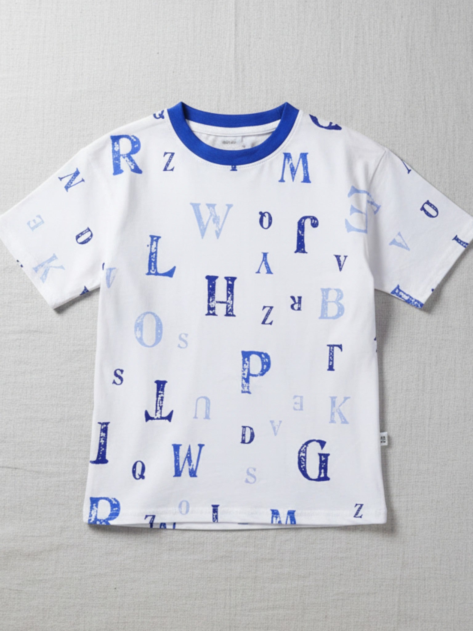 Children's white short-sleeve t-shirt with a blue crew neck, featuring a scattered print of various alphabet letters in shades of blue.