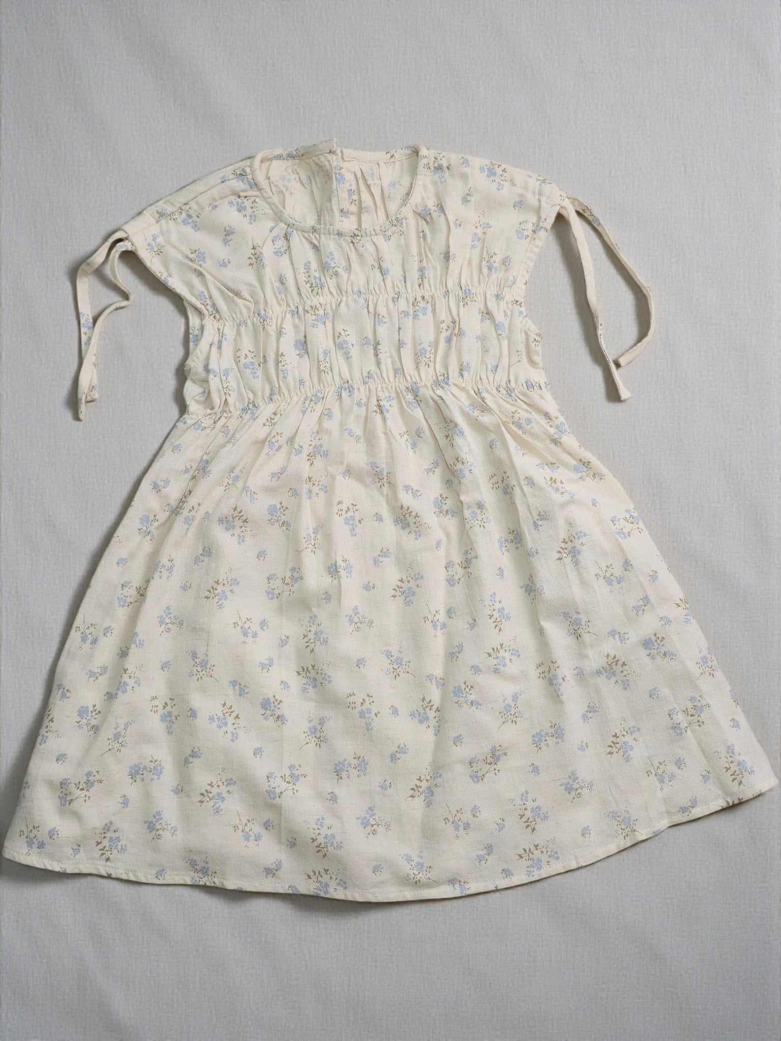 Girls short-sleeve cream dress featuring a smocked bodice, adjustable tie sleeves, and a delicate light blue floral botanical print.