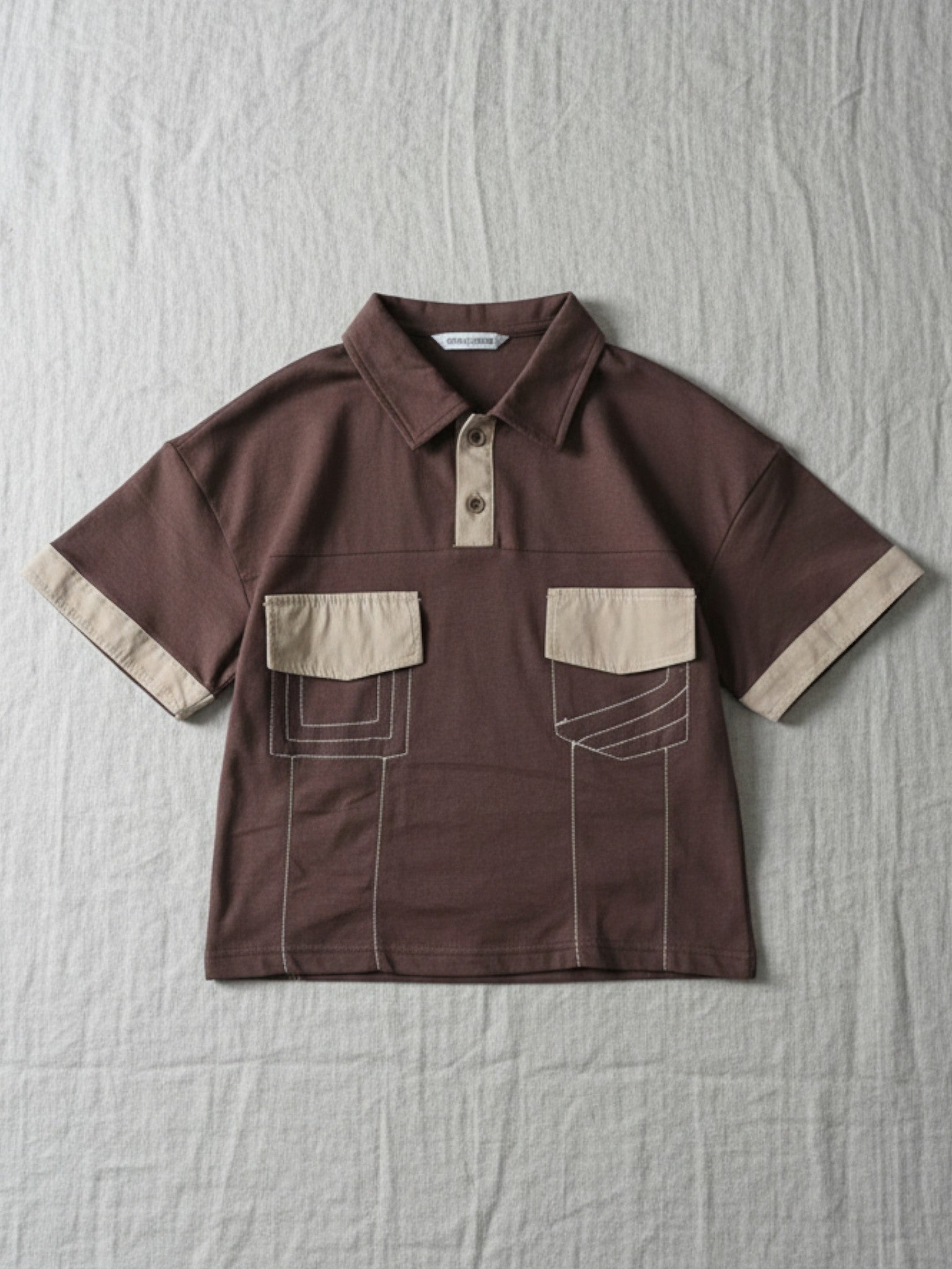 Kids short-sleeve chocolate brown polo shirt featuring contrast tan collar, cuffs, and dual chest pockets with white decorative stitching on a grey background.