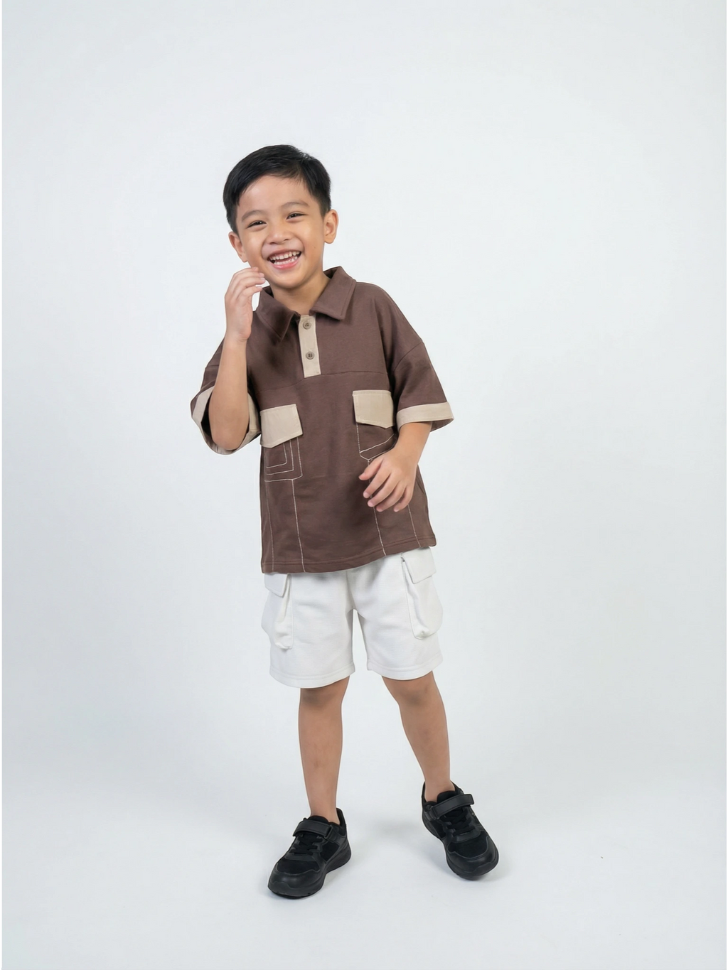Kids short-sleeve chocolate brown polo shirt featuring contrast tan collar, cuffs, and dual chest pockets with white decorative stitching on a grey background.