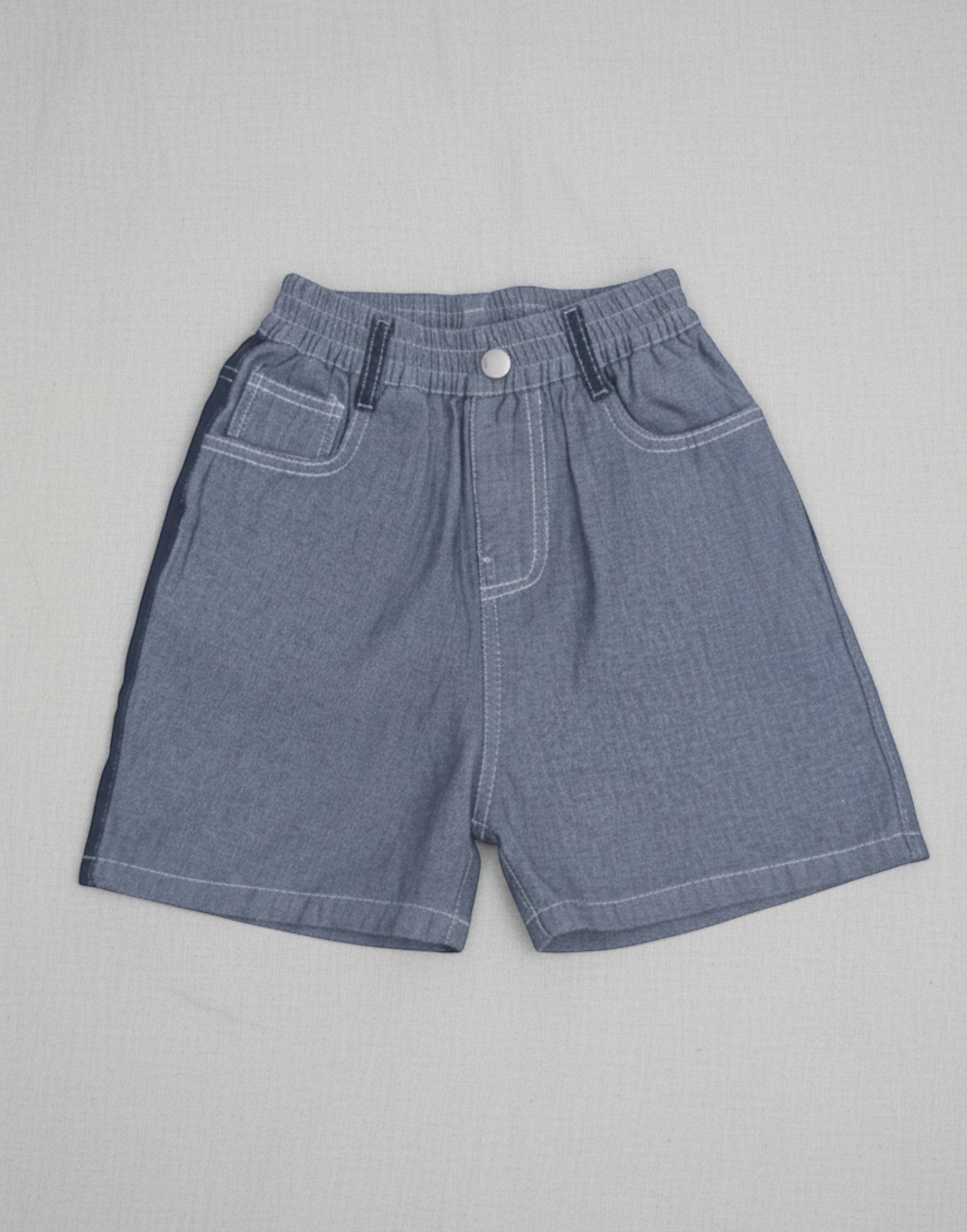 Kids blue denim shorts featuring bold white contrast stitching, a gathered elastic waistband, and a front metal button on a neutral grey background.