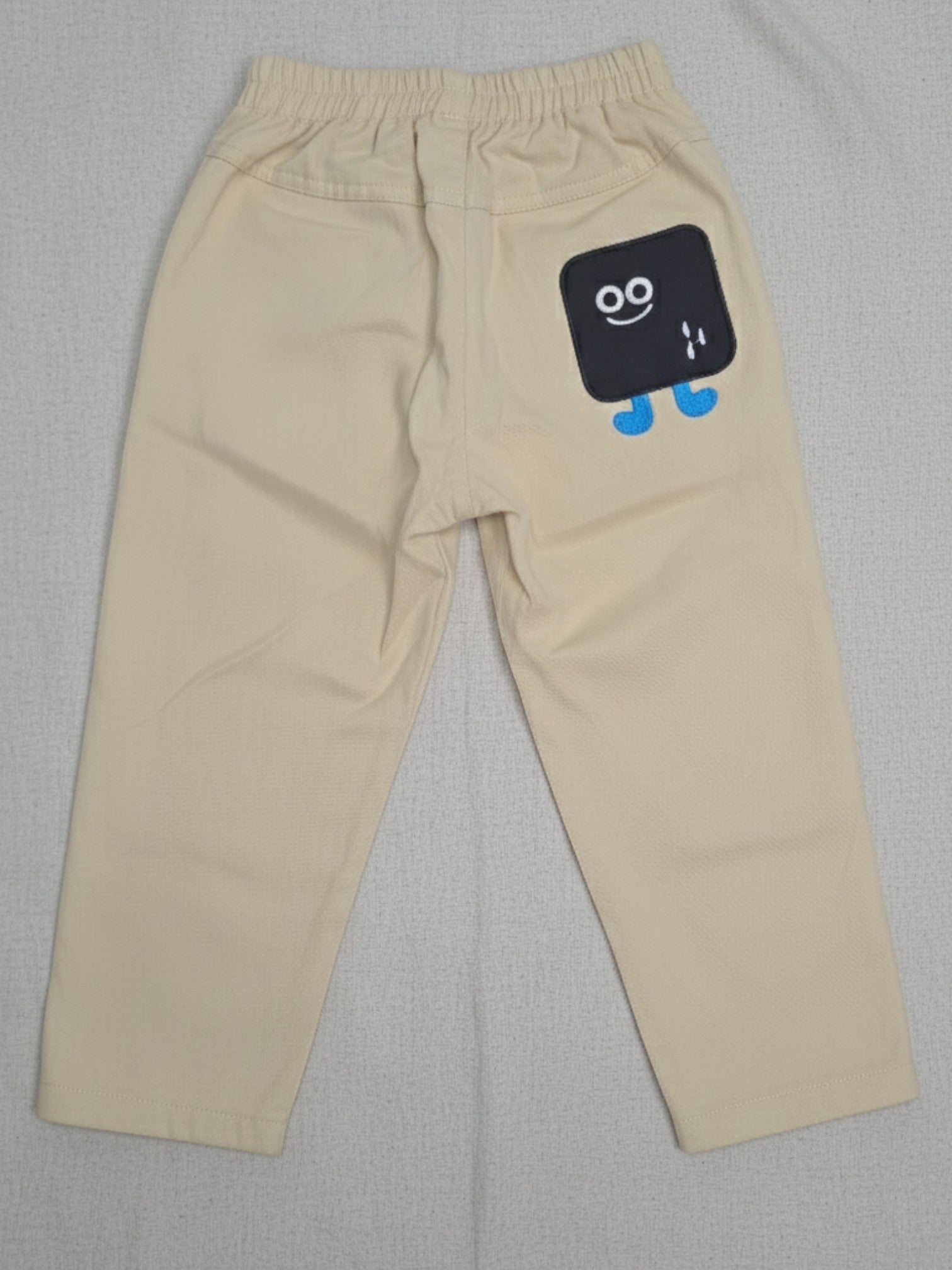 Kids cream-colored cotton trousers featuring an elasticated waistband, a front button, and a small embroidered square character on the left leg.