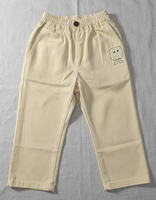 Kids cream-colored cotton trousers featuring an elasticated waistband, a front button, and a small embroidered square character on the left leg.