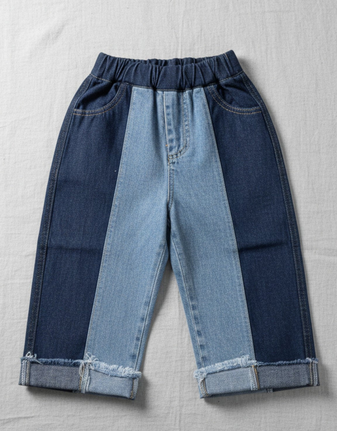 Kids blue denim jeans featuring a two-tone light and dark patchwork design, a gathered elastic waistband, and raw frayed hems on a white textured background.