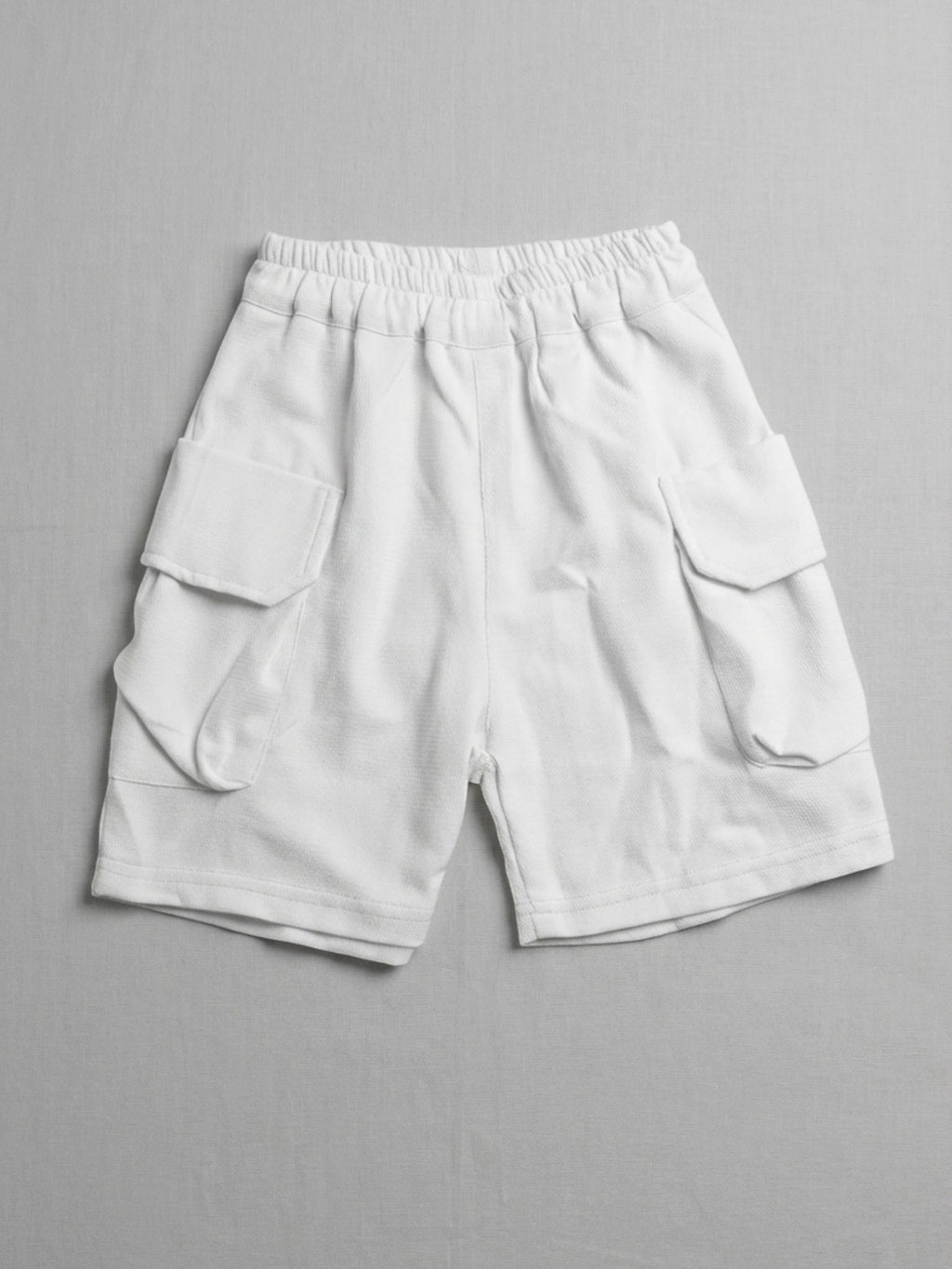Kids white short-sleeve cargo shorts with an elasticated waistband and large side flap pockets displayed on a neutral textured background.