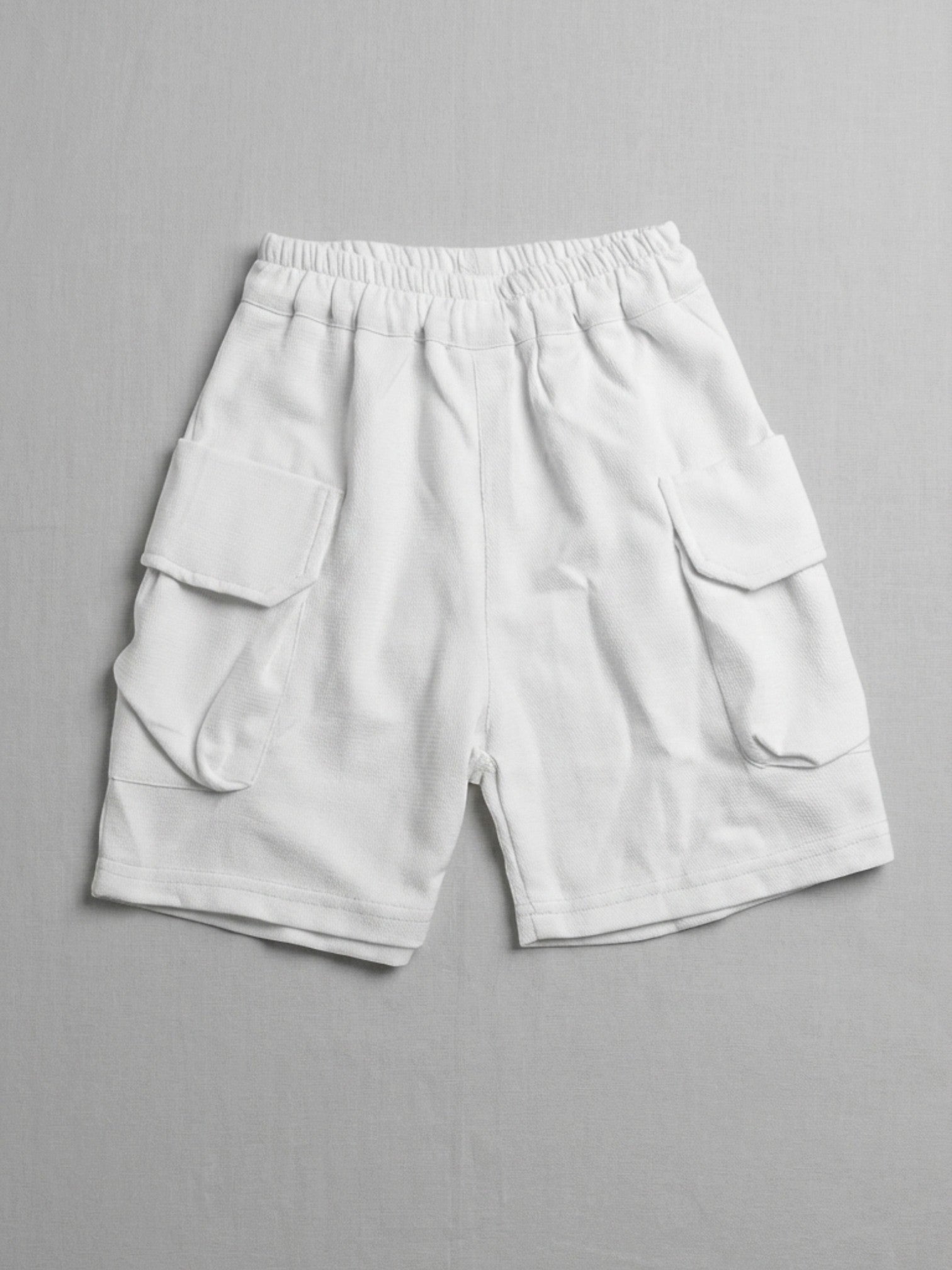 Kids white short-sleeve cargo shorts with an elasticated waistband and large side flap pockets displayed on a neutral textured background.