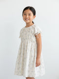 Girls short-sleeve cream dress featuring a smocked bodice, adjustable tie sleeves, and a delicate light blue floral botanical print.