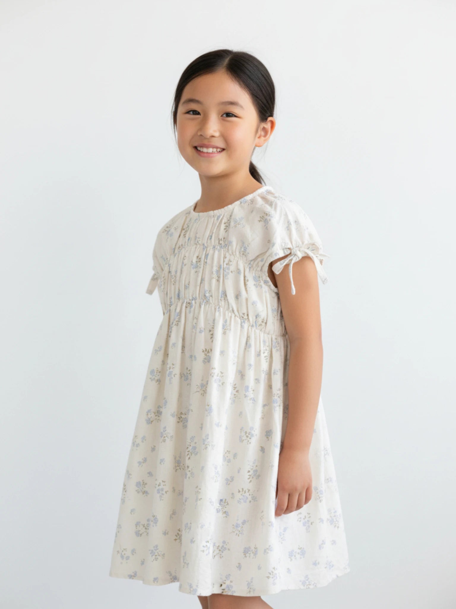 Girls short-sleeve cream dress featuring a smocked bodice, adjustable tie sleeves, and a delicate light blue floral botanical print.