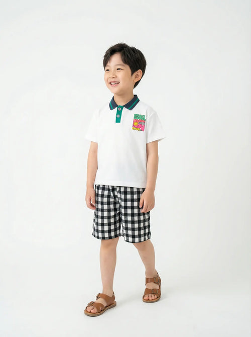 Child wearing a white polo shirt with a colorful patch and black checkered shorts on a white background