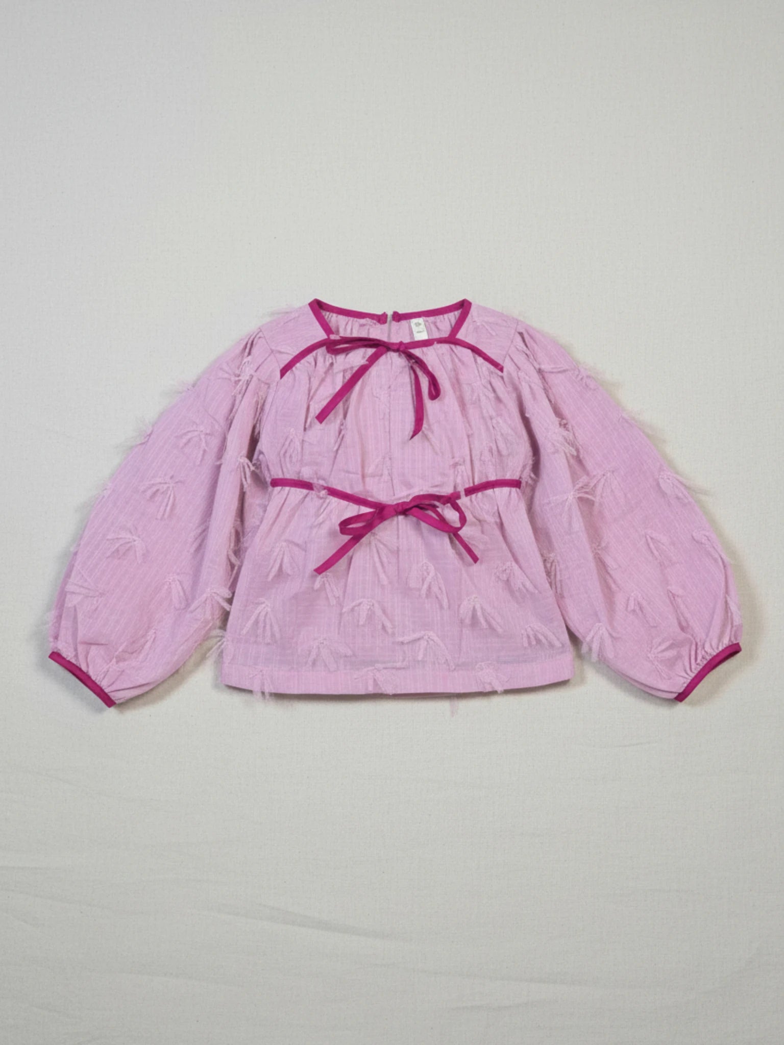 Girls' soft pink long-sleeve blouse featuring a unique 3D feathered texture, dark pink ribbon bows at the neck and waist, and elasticated cuffs.