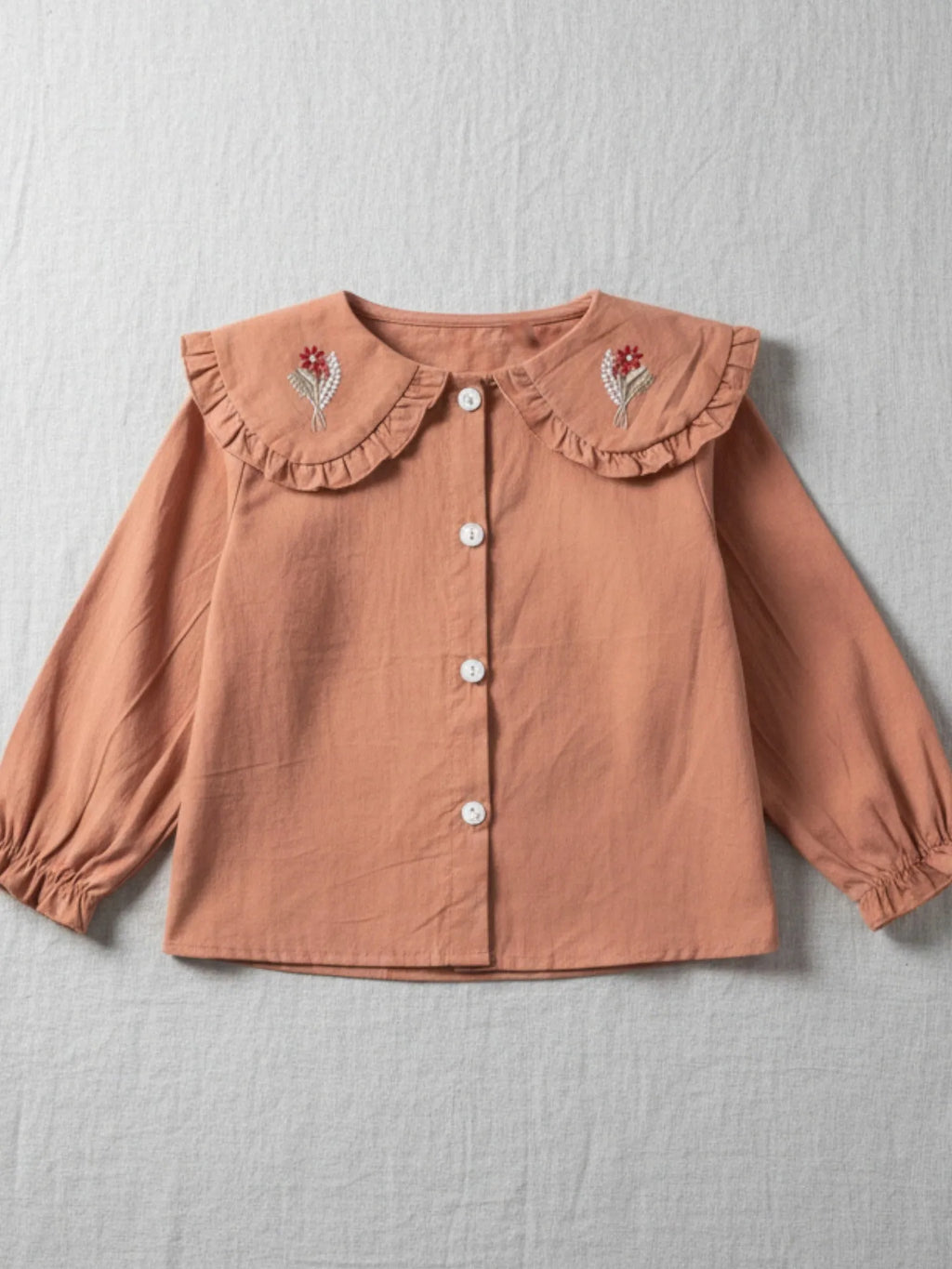 Tan long-sleeve blouse for girls featuring a ruffle-trimmed collar with pink and green floral embroidery and elasticated cuffs.