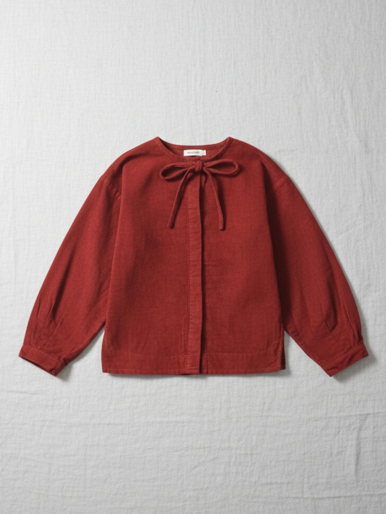 Children's long-sleeve top in a deep red corduroy fabric featuring a round neckline with a matching corduroy bow tie and hidden button placket