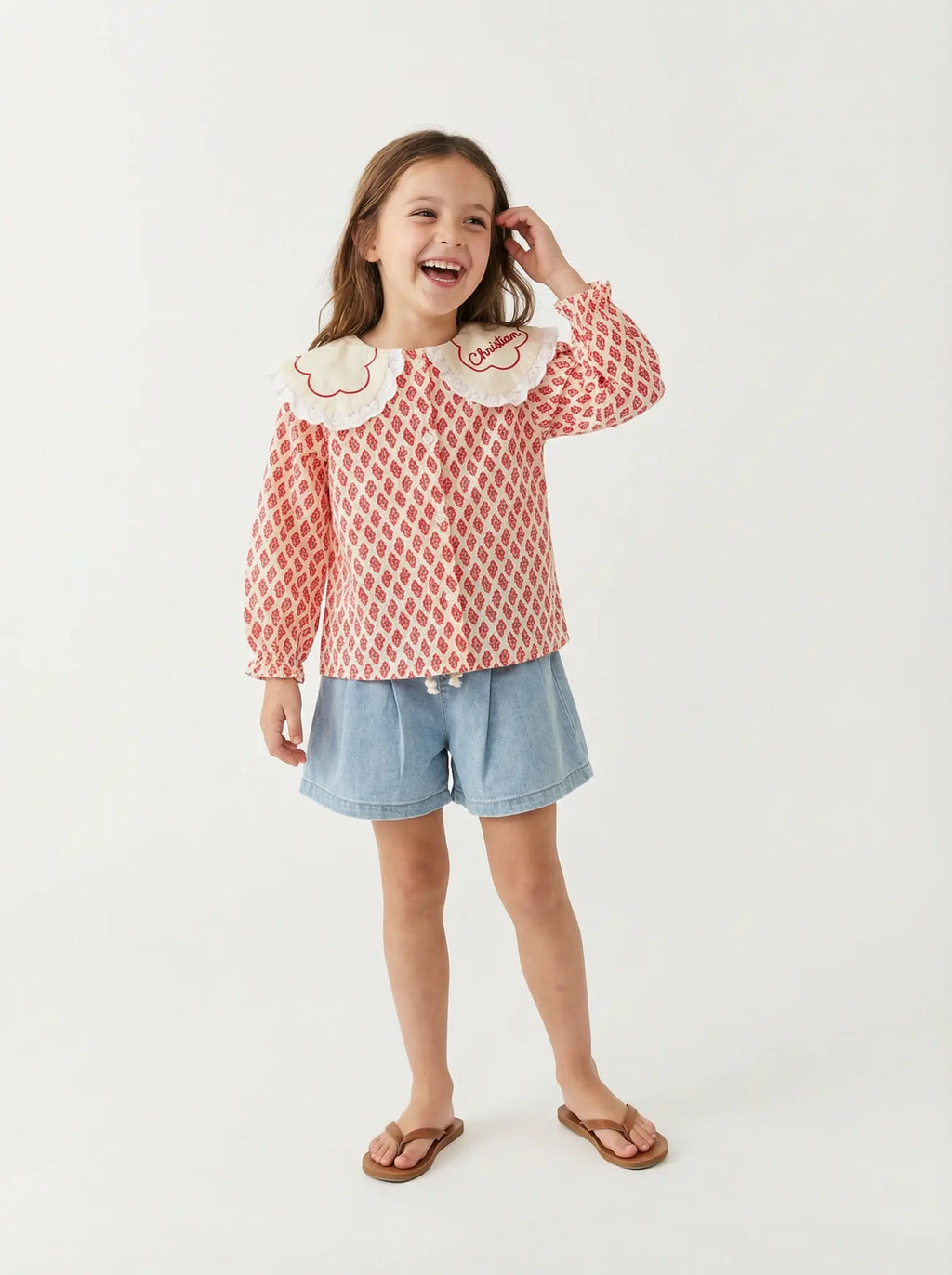 Young girl wearing a red patterned blouse with white collar and denim shorts on a white background
